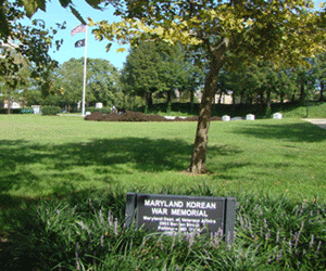 Image of the Maryland Korean War Memorial Plaque in foreground with US Flag & POW Flag in background amongst the lovely trees & gardens of Canton Waterfront Park on Boston Street, Baltimore MD