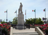 Image of Columbus Piazza statue surrounded by beautiful red flowers & colorful waving Italian flags on President Street at Eastern Avenue, Baltimore MD